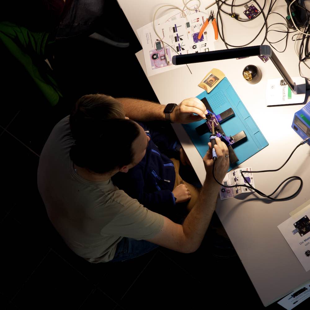 Zwei Personen sitzen an einem Tisch und arbeiten gemeinsam an einem elektronischen Nachtlicht-Bausatz. Sie konzentrieren sich auf die Montage einer Platine, umgeben von verschiedenen Elektronikwerkzeugen und Informationsmaterialien.