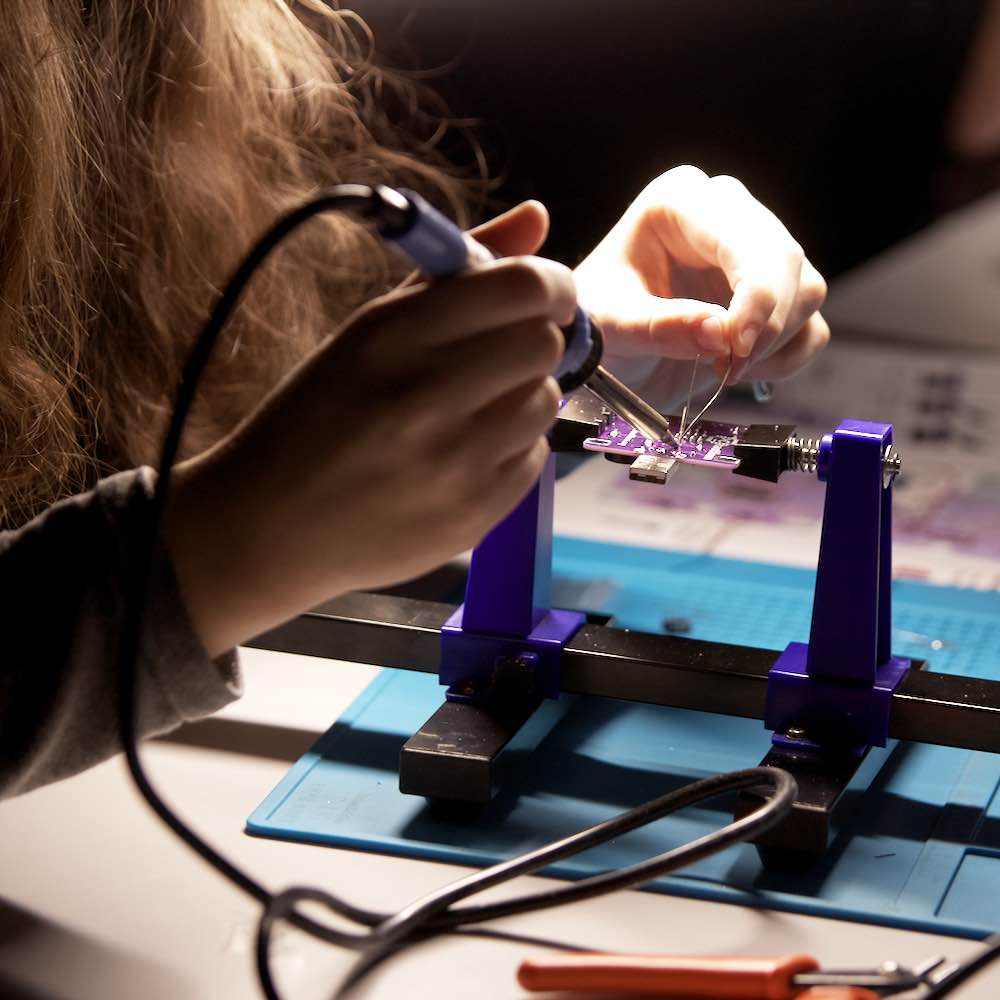 Ein Kind arbeitet an einer violett gefärbten Leiterplatte, die in einem blauen PCB-Halter befestigt ist. Der Fokus liegt auf den kleinen Händen des Kindes, die einen Lötkolben halten und an der Elektronik arbeiten, mit unscharfem Werkzeug im Vordergrund und einer beleuchteten Arbeitsfläche.