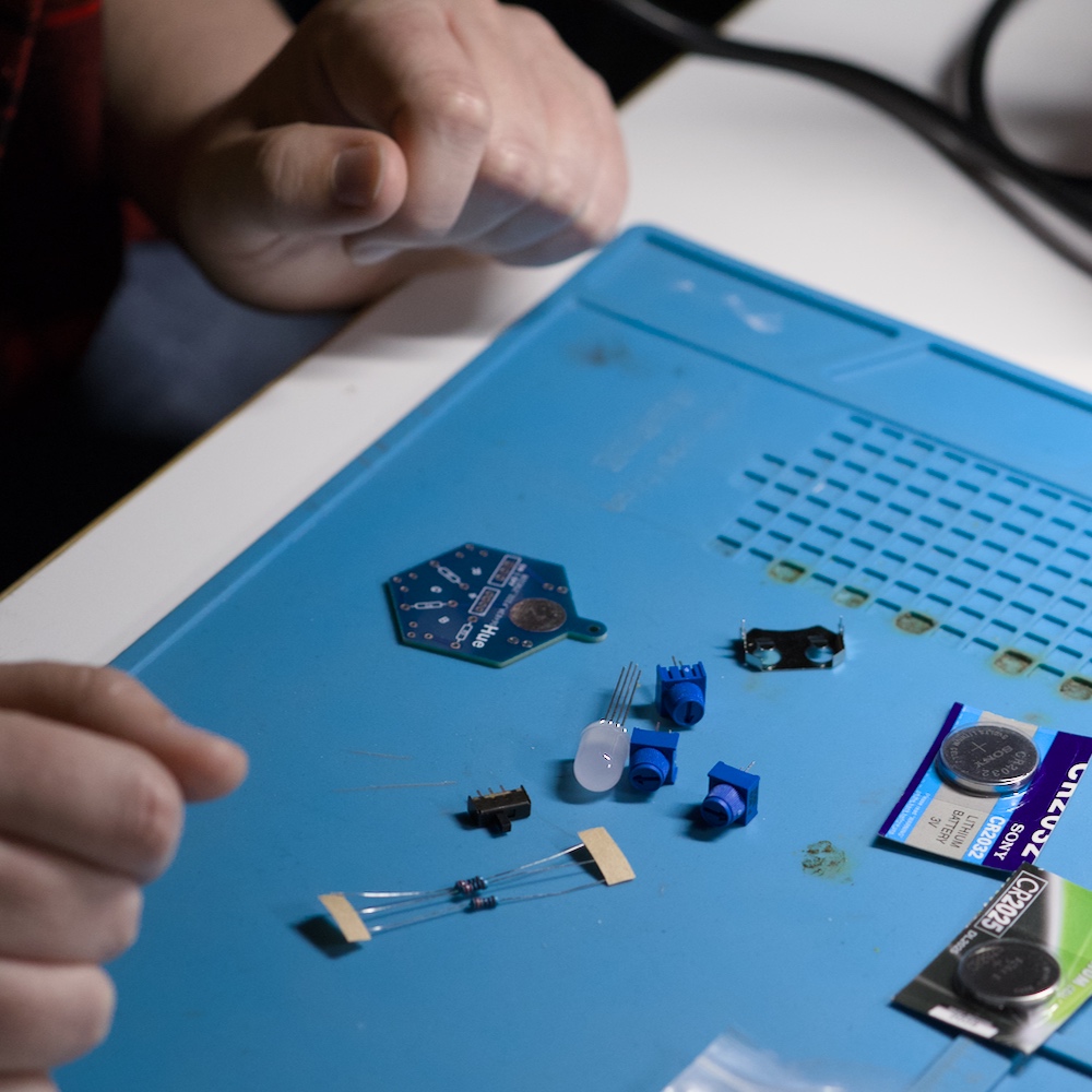 Eine kleine Leiterplatte und verschiedene Elektronikbauteile – Widerstände, LED, Taster/Trimmpotis, Steckverbinder und ein Knopfzellenhalter – liegen auf einer blauen Arbeitsmatte, umgeben von zwei Händen.