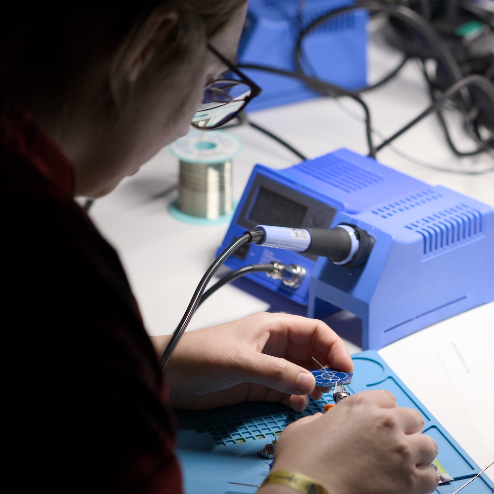 Eine Person lötet an einer kleinen Leiterplatte; vor ihr stehen eine blaue Lötstation und eine Spule Lötzinn auf einem hellen Arbeitstisch.