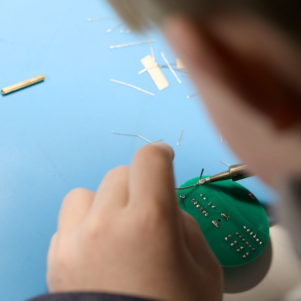 Eine Person lötet an der Unterseite einer grünen Leiterplatte; auf der blauen Arbeitsmatte liegen abgeschnittene Drahtreste.