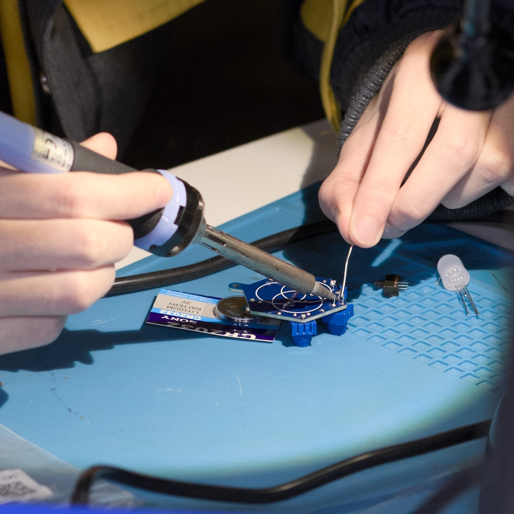 Eine Person lötet an einer kleinen blauen Leiterplatte und führt dabei Lötzinn an die Lötspitze; daneben liegen weitere elektronische Bauteile und eine Knopfzellenverpackung.