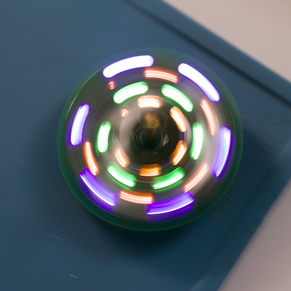 Ein rotierender Kreisel mit mehrfarbigen LEDs erzeugt leuchtende, ringförmige Lichtspuren auf einer blauen Unterlage.