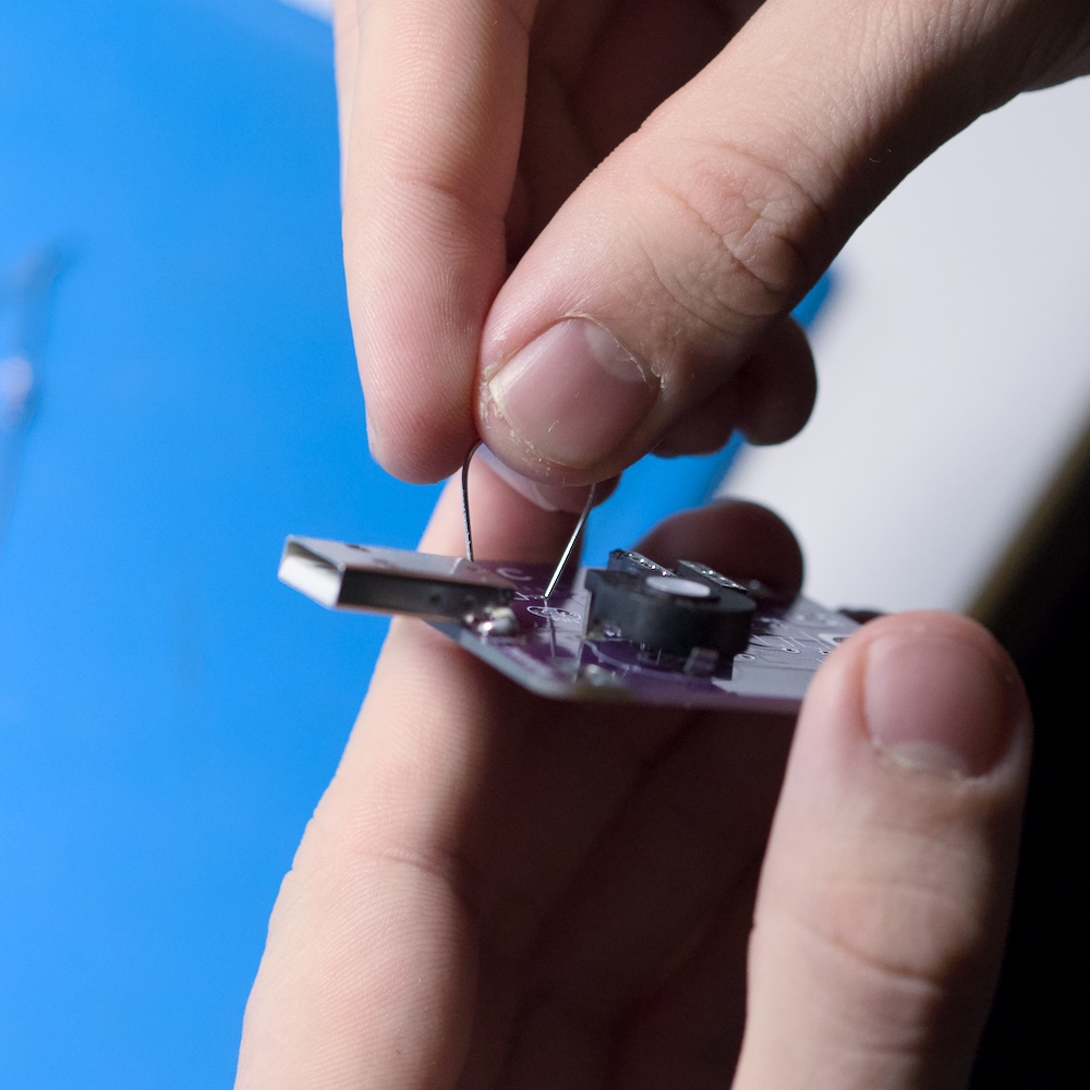 Zwei Hände stecken die Drahtanschlüsse eines Bauteils in eine kleine Platine mit USB-Stecker und montiertem Potentiometer.