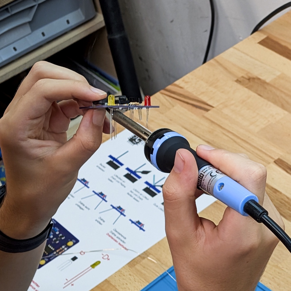 Zwei Hände halten eine schmale Leiterplatte mit roten und gelben LEDs, während ein Lötkolben an die herausstehenden Anschlussdrähte geführt wird; darunter liegt eine bebilderte Bauanleitung.