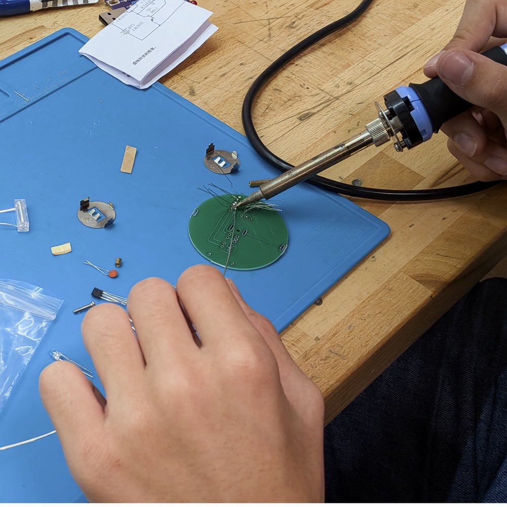 Eine Person lötet Anschlussdrähte auf der Unterseite einer runden grünen Platine; auf der blauen Arbeitsmatte liegen zwei Knopfzellenhalter und weitere Bauteile.