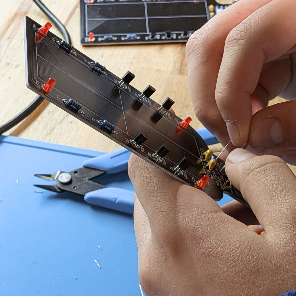 Zwei Hände führen die Anschlussdrähte eines Bauteils durch eine längliche schwarze Leiterplatte, die bereits mit roten LEDs, Widerständen und Transistoren bestückt ist.