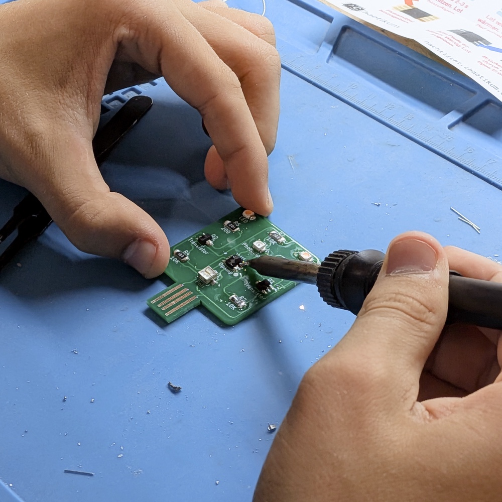 Eine Person lötet mit feiner Lötspitze an einer grünen SMD-Nachtlicht-Platine; auf der Platine sind unter anderem Photodiode, OpAmp und Transistor beschriftet.